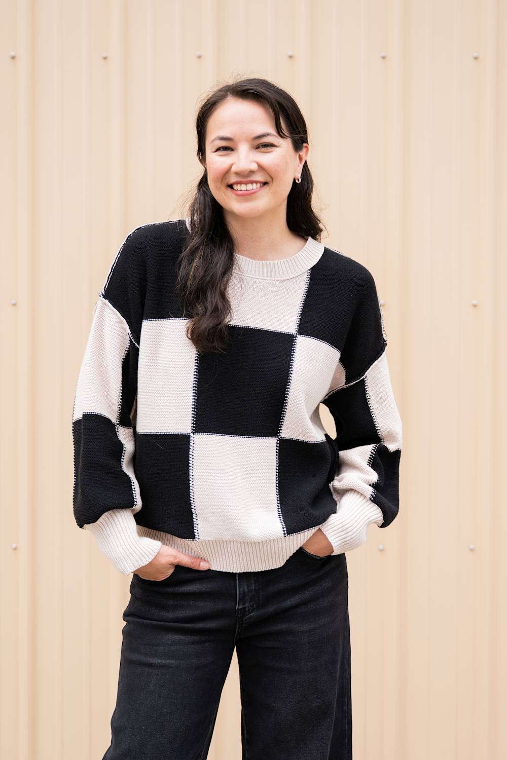 A woman wearing a black and off-white checkered knit sweater and dark jeans, standing against a beige corrugated metal wall.