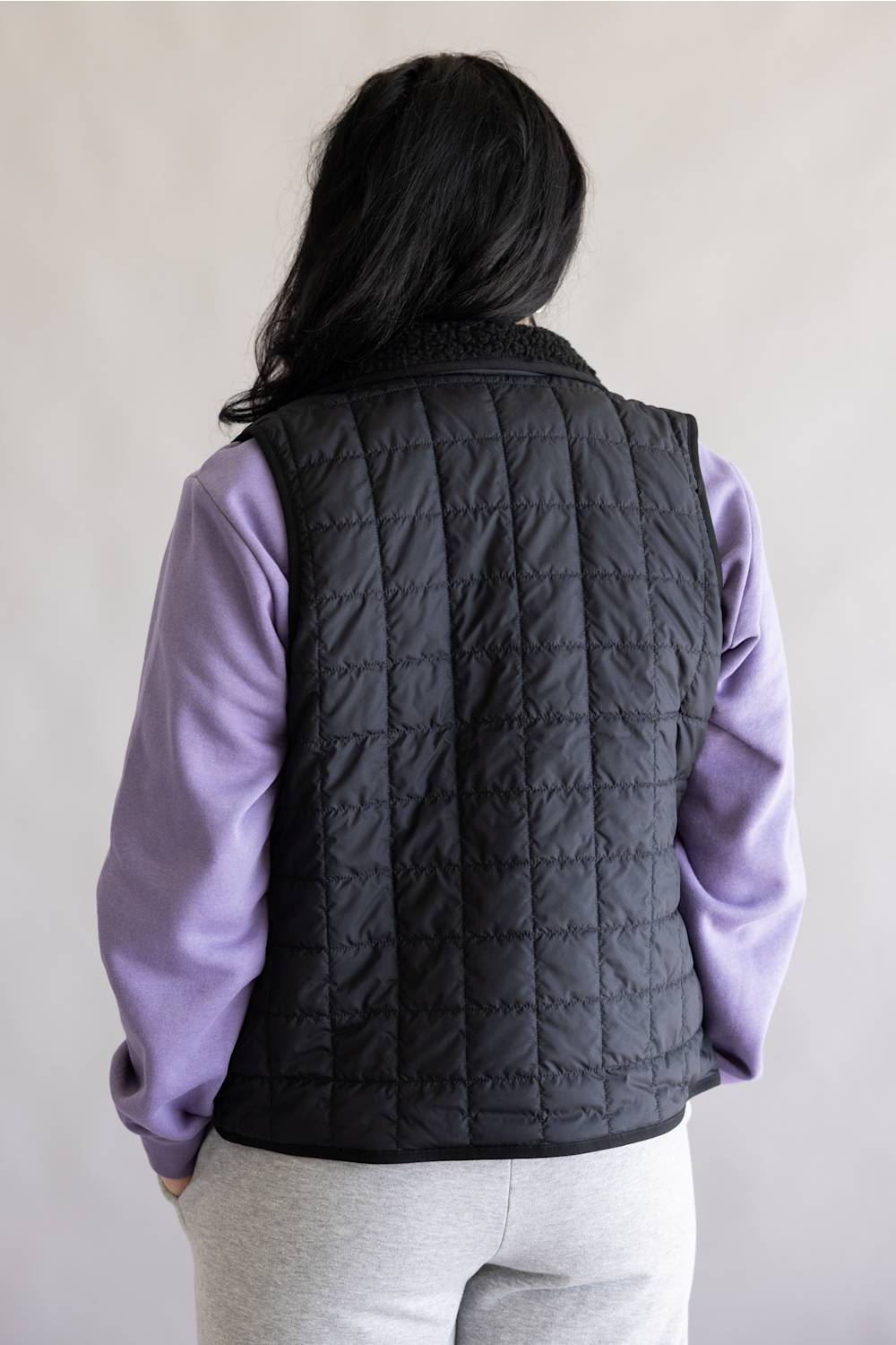 A person shown from the back wearing a black quilted puffer vest over a lavender long-sleeve top and light gray sweatpants, against a plain light gray background.