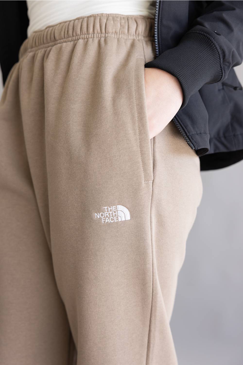 Close-up of a person wearing beige jogger sweatpants with a white embroidered The North Face logo on the left thigh, paired with a dark jacket.