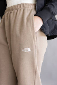 Close-up of a person wearing beige jogger sweatpants with a white embroidered The North Face logo on the left thigh, paired with a dark jacket.