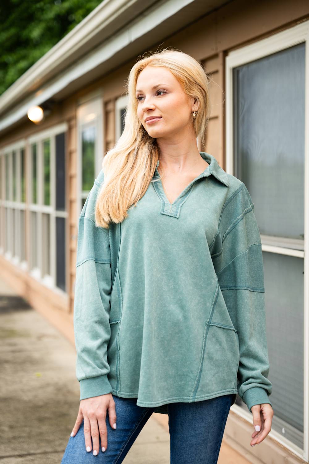 A model stands outdoors wearing a faded sage green oversized pullover with a collared neckline and dark blue jeans in front of a building with large windows.