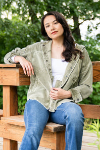 A woman sitting on a wooden bench outdoors, wearing an olive green corduroy button-up shirt over a white tank top and blue jeans, with a blurred green garden background.