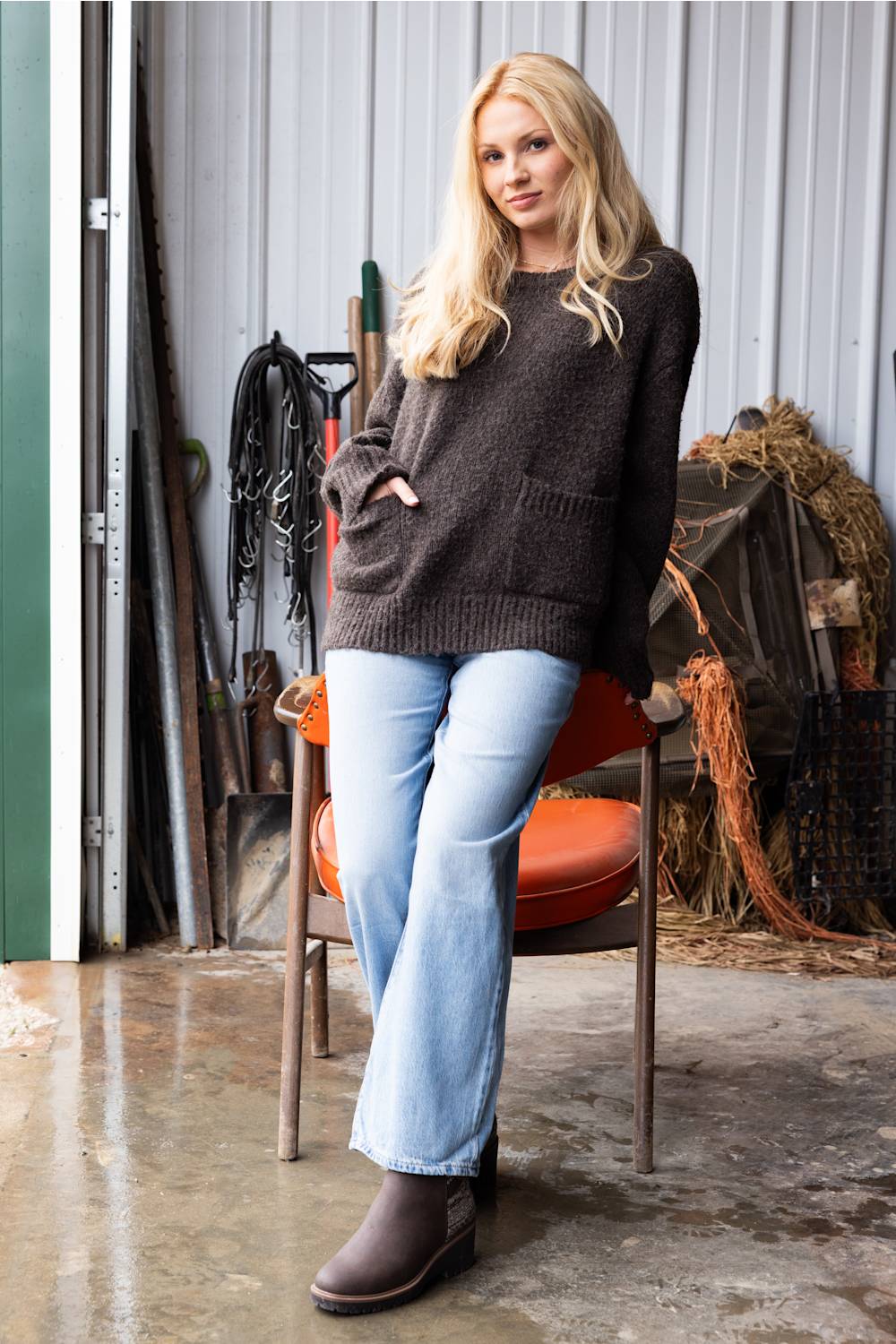 A woman stands in a rustic barn-like setting wearing a dark charcoal knit sweater, light blue flare jeans, and brown ankle boots, leaning against an orange chair.