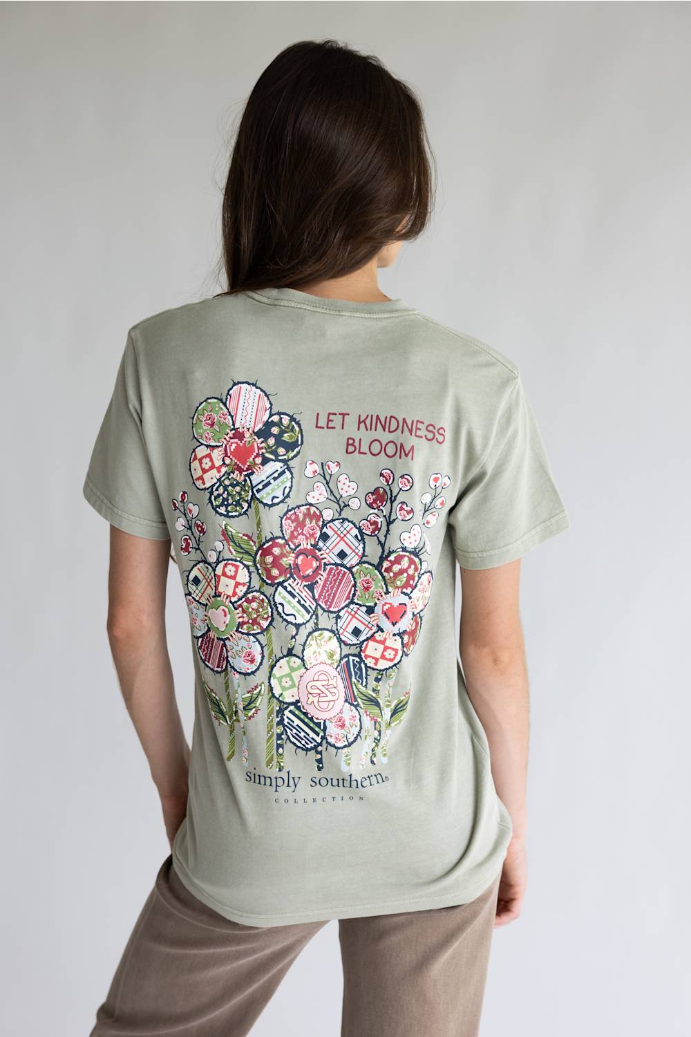 A light beige crewneck t-shirt with a colorful floral graphic on the back, featuring the text 'LET KINDNESS BLOOM' and 'simply southern.' COLLECTIONS.