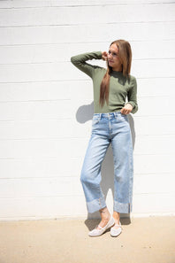 Woman posing against a white painted brick wall wearing a long-sleeve olive green crewneck shirt, cuffed light-wash blue jeans, and white woven flats.