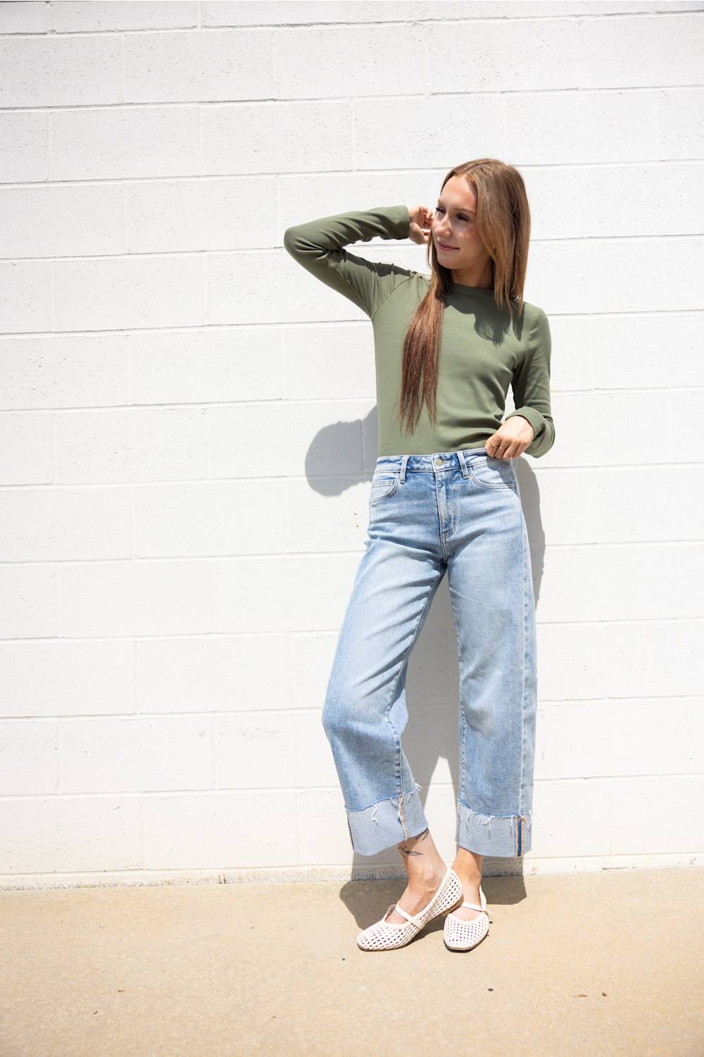 Woman posing against a white painted brick wall wearing a long-sleeve olive green crewneck shirt, cuffed light-wash blue jeans, and white woven flats.