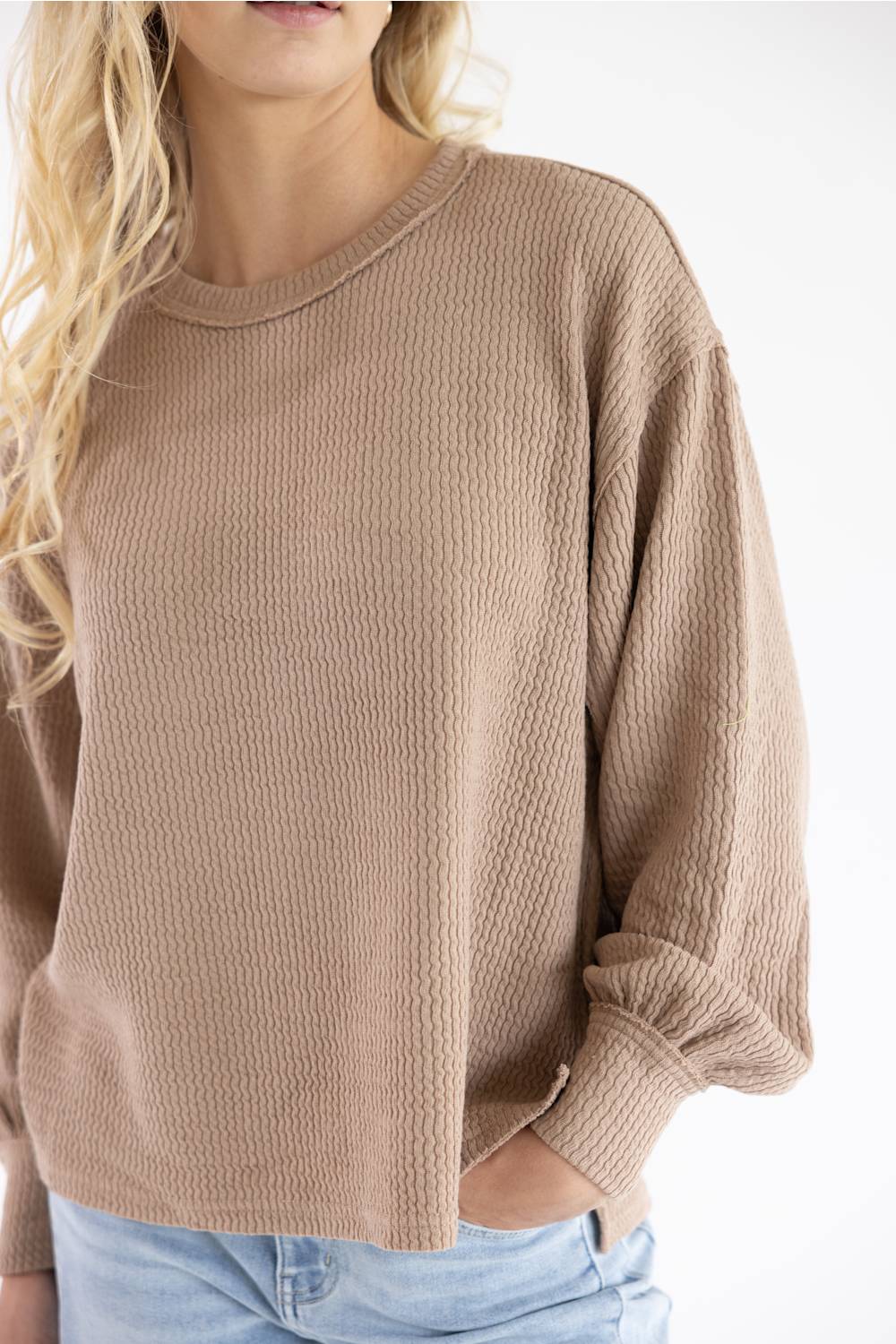 A cropped close-up of a woman's torso wearing a light brown, waffle-knit long-sleeve sweater, showing the texture, the round neckline, the ribbed cuffs, and her hand resting in the front pocket. She also wears light blue jeans. Her blonde hair is visible on the left side of the frame. The background is a plain white wall.