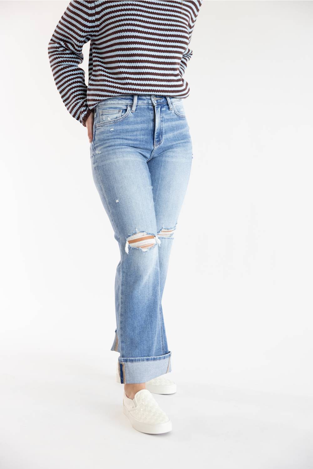 A person wearing light-wash blue distressed jeans with rolled cuffs, paired with a brown and blue striped long-sleeve top and white slip-on shoes, photographed against a plain white studio background.