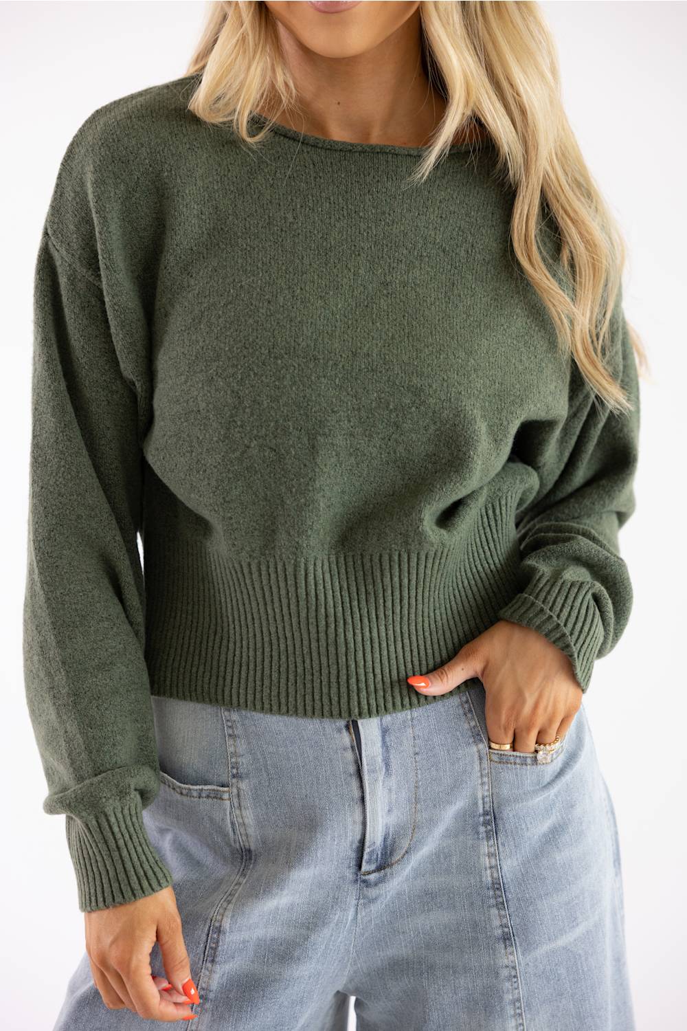 Torso-level view of a model wearing an olive green knit boat neck sweater paired with light wash denim jeans, hand tucked in pocket.
