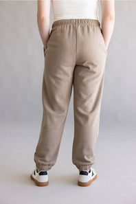 A person shown from the back wearing loose beige jogger sweatpants with an elastic waistband and cuffs, paired with white sneakers, against a plain light gray studio background.