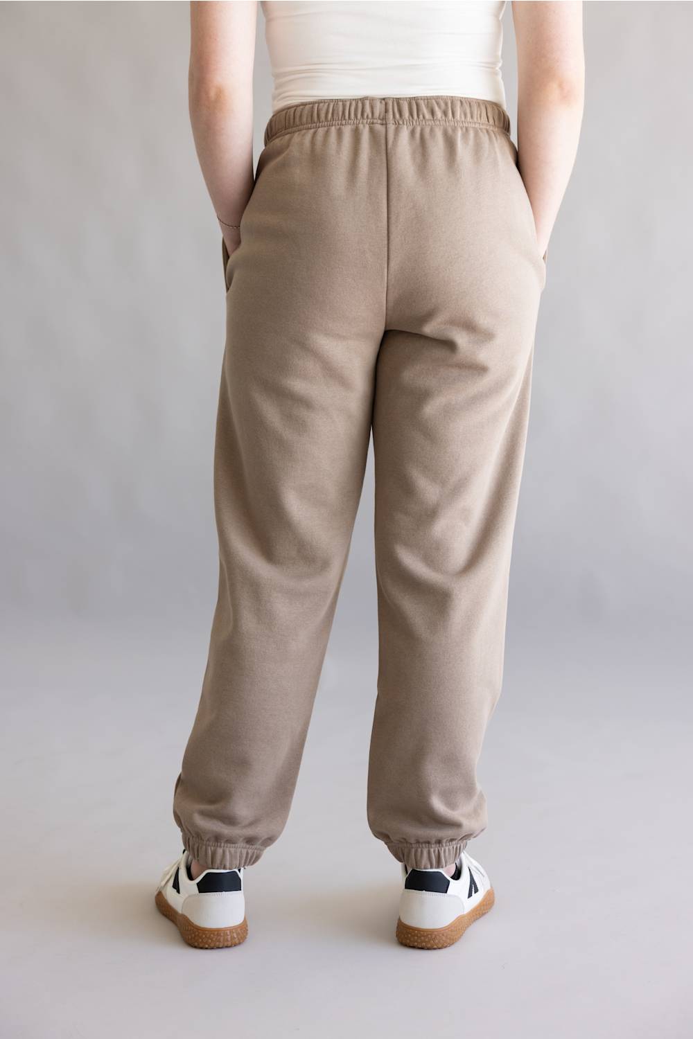 A person shown from the back wearing loose beige jogger sweatpants with an elastic waistband and cuffs, paired with white sneakers, against a plain light gray studio background.