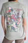 A light beige crewneck t-shirt with a colorful floral graphic on the back, featuring the text 'LET KINDNESS BLOOM' and 'simply southern.' COLLECTIONS.