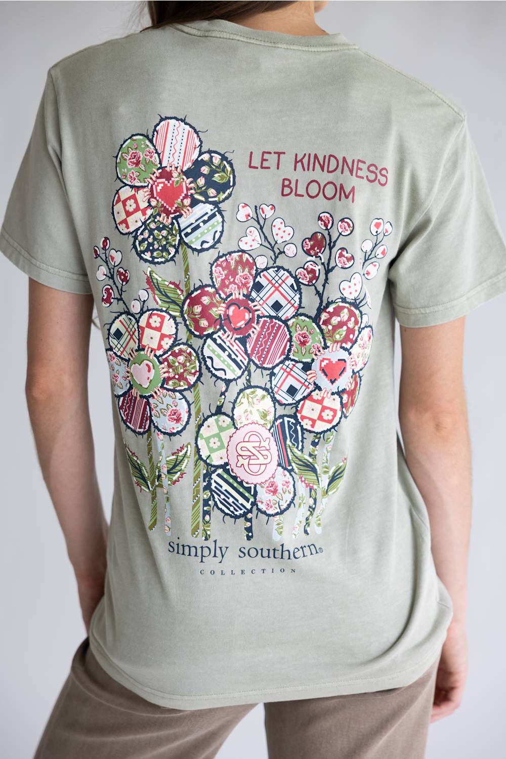 A light beige crewneck t-shirt with a colorful floral graphic on the back, featuring the text 'LET KINDNESS BLOOM' and 'simply southern.' COLLECTIONS.