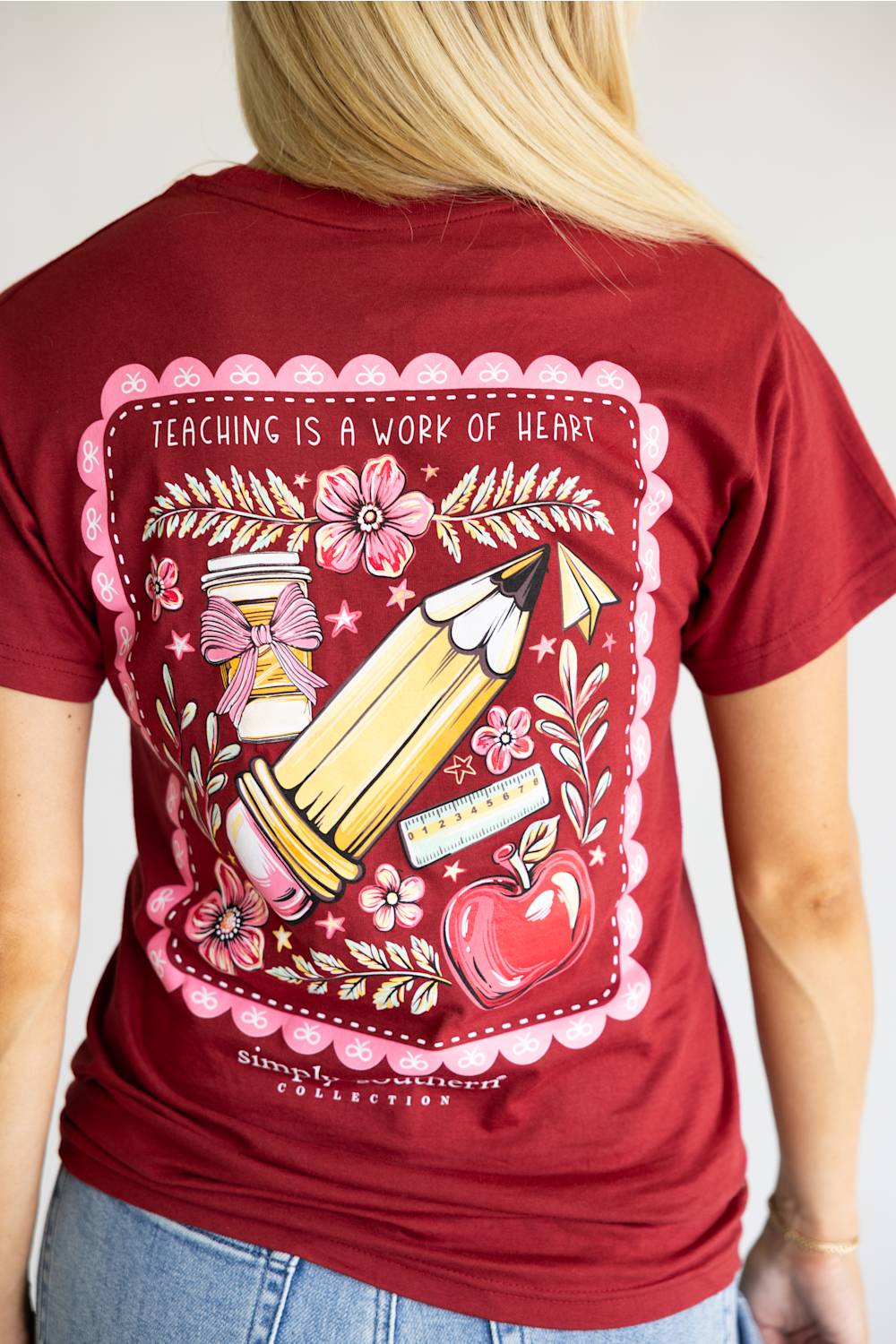 A red short-sleeve t-shirt with a large graphic print on the back, featuring a pink scalloped border, a yellow pencil, a red apple, and the text 'TEACHING IS A WORK OF ART' and 'simply southern' below it.