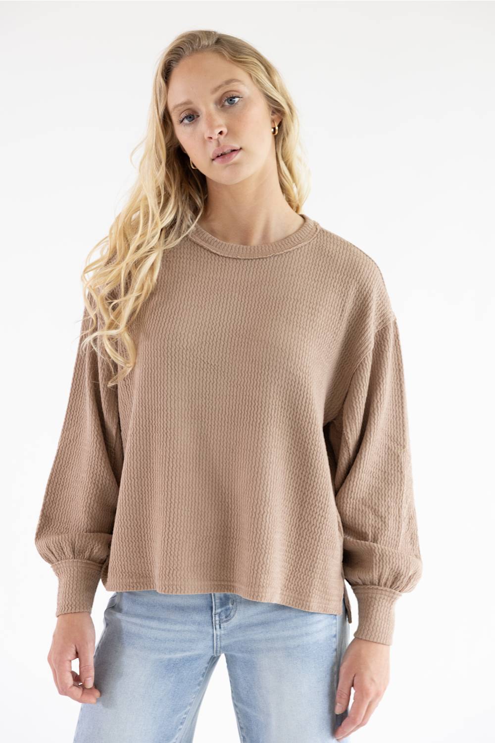 Front view of a blonde woman wearing a khaki waffle-knit, crewneck sweater with relaxed long sleeves, tight ribbed cuffs, and light blue jeans against a plain white background.