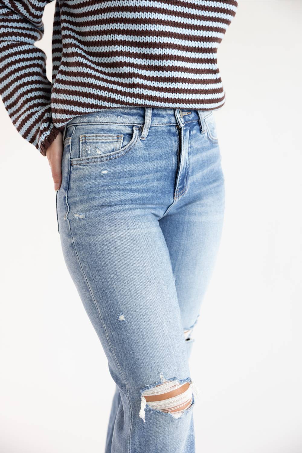 A person wearing light-wash blue distressed jeans with ripped details at the knees, paired with a brown and blue striped knit top, shown against a plain white background.