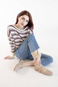 A seated model in a white studio setting, wearing a striped purple and beige knit sweater, light wash frayed denim jeans, and beige perforated knit Mary Jane flats with single adjustable buckles.