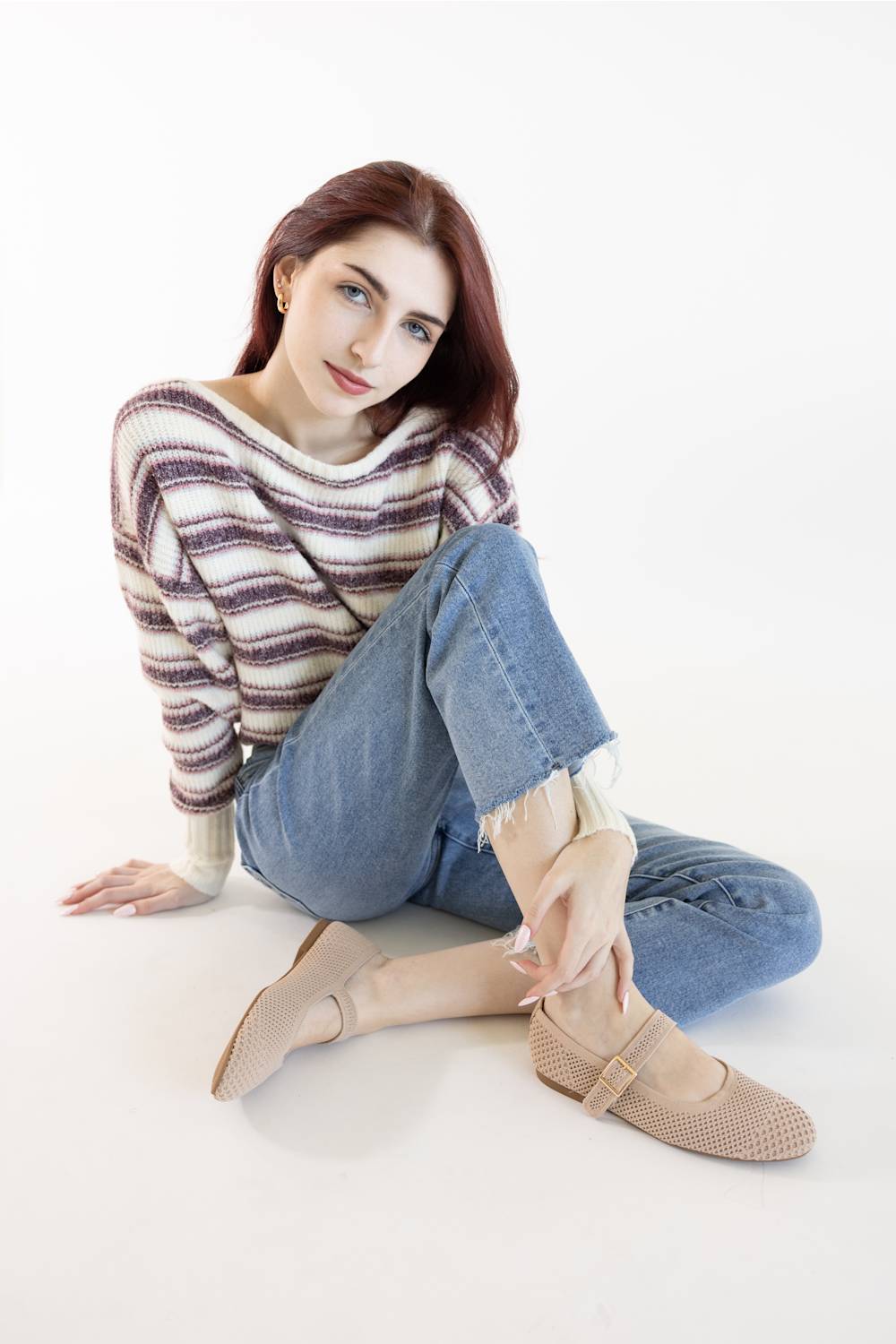 A seated model in a white studio setting, wearing a striped purple and beige knit sweater, light wash frayed denim jeans, and beige perforated knit Mary Jane flats with single adjustable buckles.