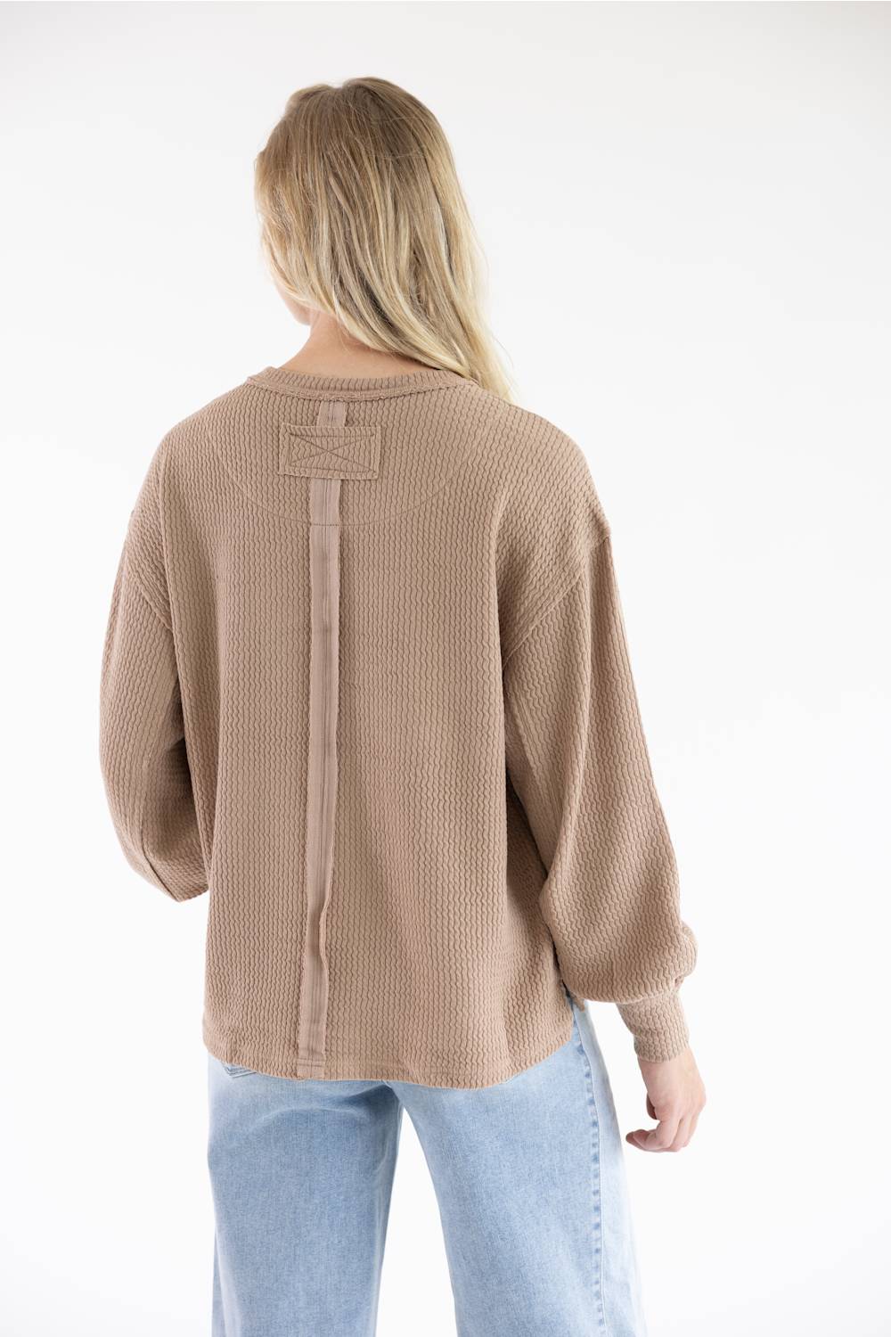 Back view of a woman with long blonde hair wearing a khaki ribbed knit, waffle-texture sweater with vertical fabric tape trim detailing down the center back and light blue jeans against a plain white background.