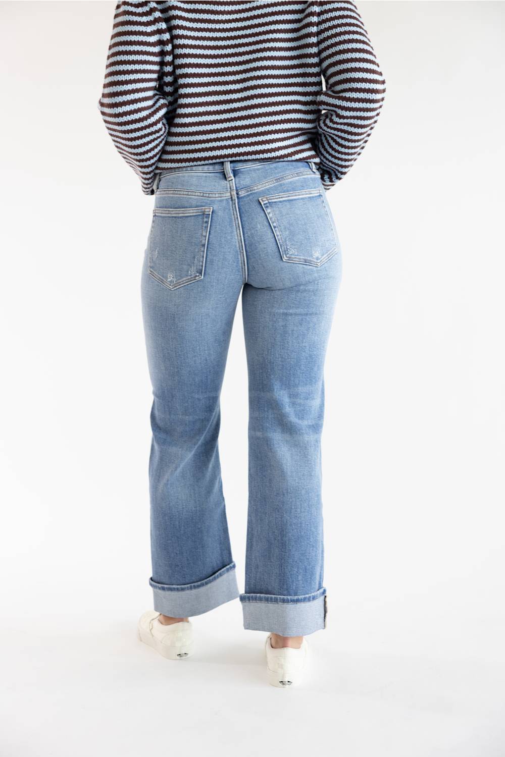 A person shown from the back wearing light-wash blue jeans with rolled cuffs, a brown and blue striped long-sleeve top, and white shoes, against a plain white studio background.