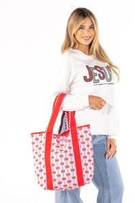 A woman modeling a white long-sleeve sweatshirt with a red and black graphic print, paired with light blue jeans and carrying a white tote bag with red handles and a red and white pattern.