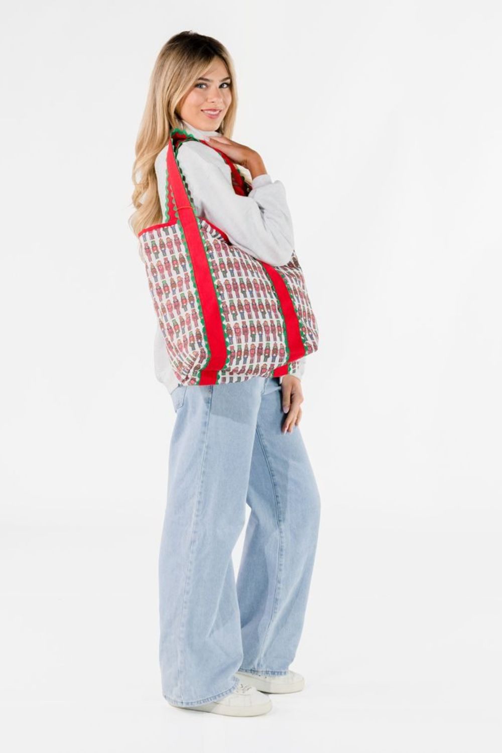 A woman modeling a large woven tote bag with a red, green, and white houndstooth pattern and solid red shoulder straps, paired with a white long-sleeve top, light blue wide-leg jeans, and white sneakers against a plain white studio background.