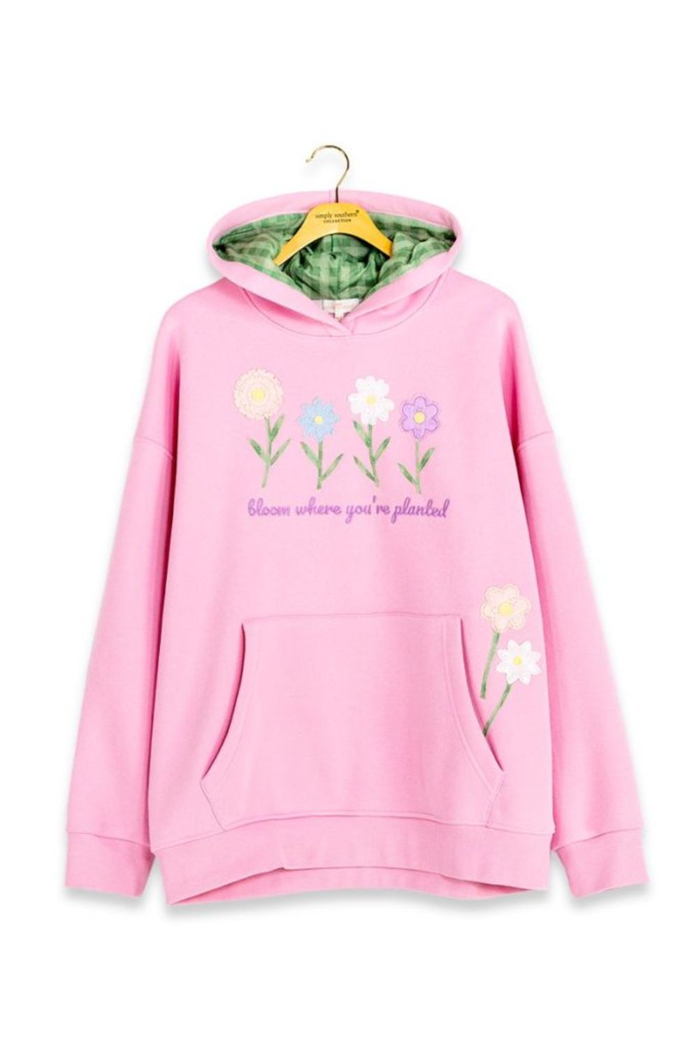 A pink pullover hoodie hanging on a wooden hanger against a white background. The hoodie features embroidered flowers and the text 'bloom where you're planted!' on the front, with a green patterned lining inside the hood.