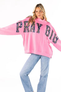A woman modeling a bright pink sweatshirt with 'PRAY BIG' written across the front in large black letters filled with a floral pattern, paired with light blue jeans against a plain white background.