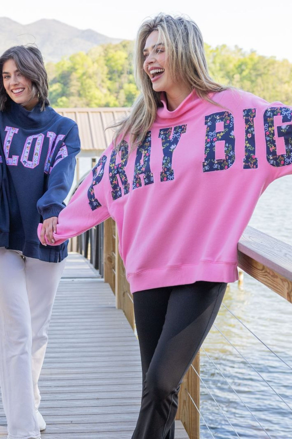 A woman in a bright pink sweatshirt with 'PRAY BIG' in glittery letters walks along a wooden dock beside water, holding hands with another woman wearing a navy sweatshirt.
