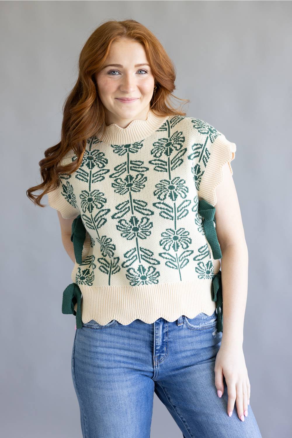 A smiling woman with long wavy red hair models a cream knit top with green floral embroidery and side ties, paired with blue jeans, against a plain gray studio background.