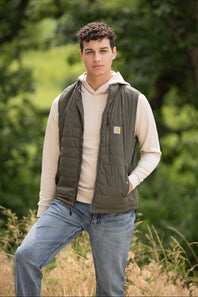 A young male model standing outdoors in a field, wearing the Carhartt Light Weight Insulated Vest for Men in Moss, layered over a cream hoodie sweatshirt and paired with light-wash blue jeans.