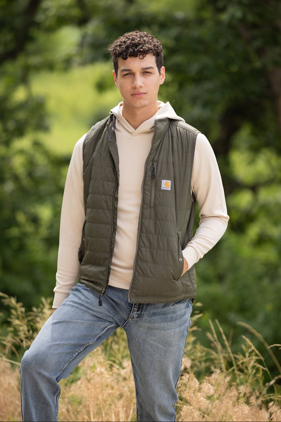 A young male model standing outdoors in a field, wearing the Carhartt Light Weight Insulated Vest for Men in Moss, layered over a cream hoodie sweatshirt and paired with light-wash blue jeans.