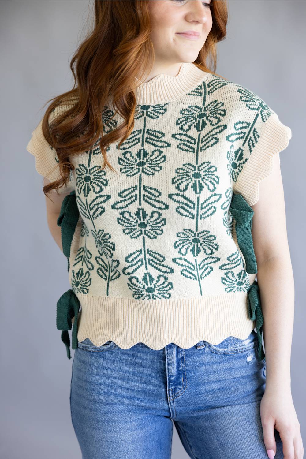 A smiling woman with long wavy red hair models a cream knit top with green floral embroidery and side ties, paired with blue jeans, against a plain gray studio background.
