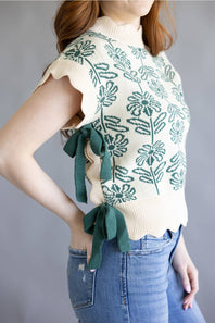 A woman wearing a cream knit sweater vest with a green floral pattern and matching green side ties, paired with blue jeans against a gray background.