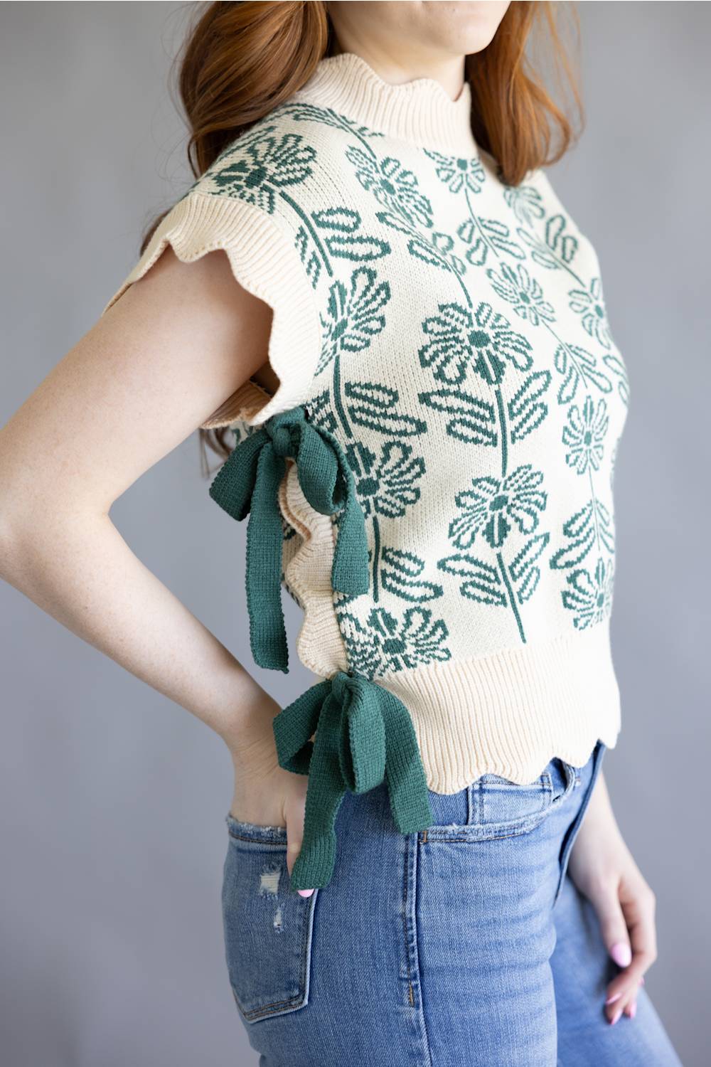 A woman wearing a cream knit sweater vest with a green floral pattern and matching green side ties, paired with blue jeans against a gray background.