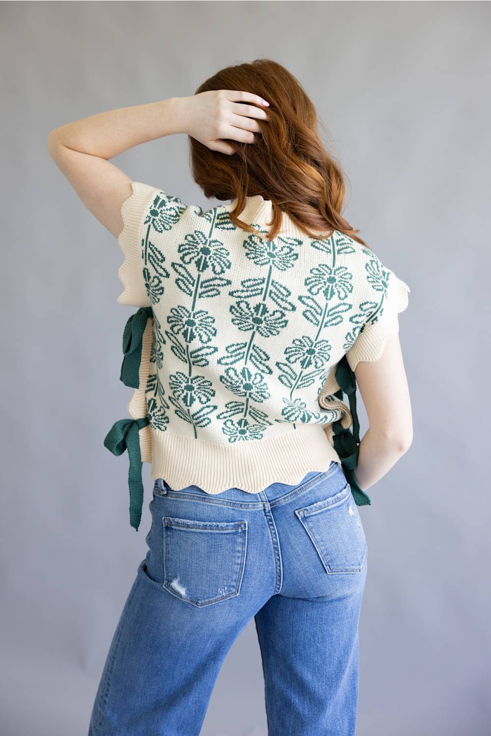 A woman shown from the back wearing a cream knit top with a green floral pattern and side ties, paired with blue jeans, against a plain gray studio background.