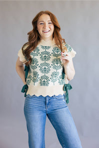 A smiling woman with long wavy red hair models a cream knit top with green floral embroidery and side ties, paired with blue jeans, against a plain gray studio background.