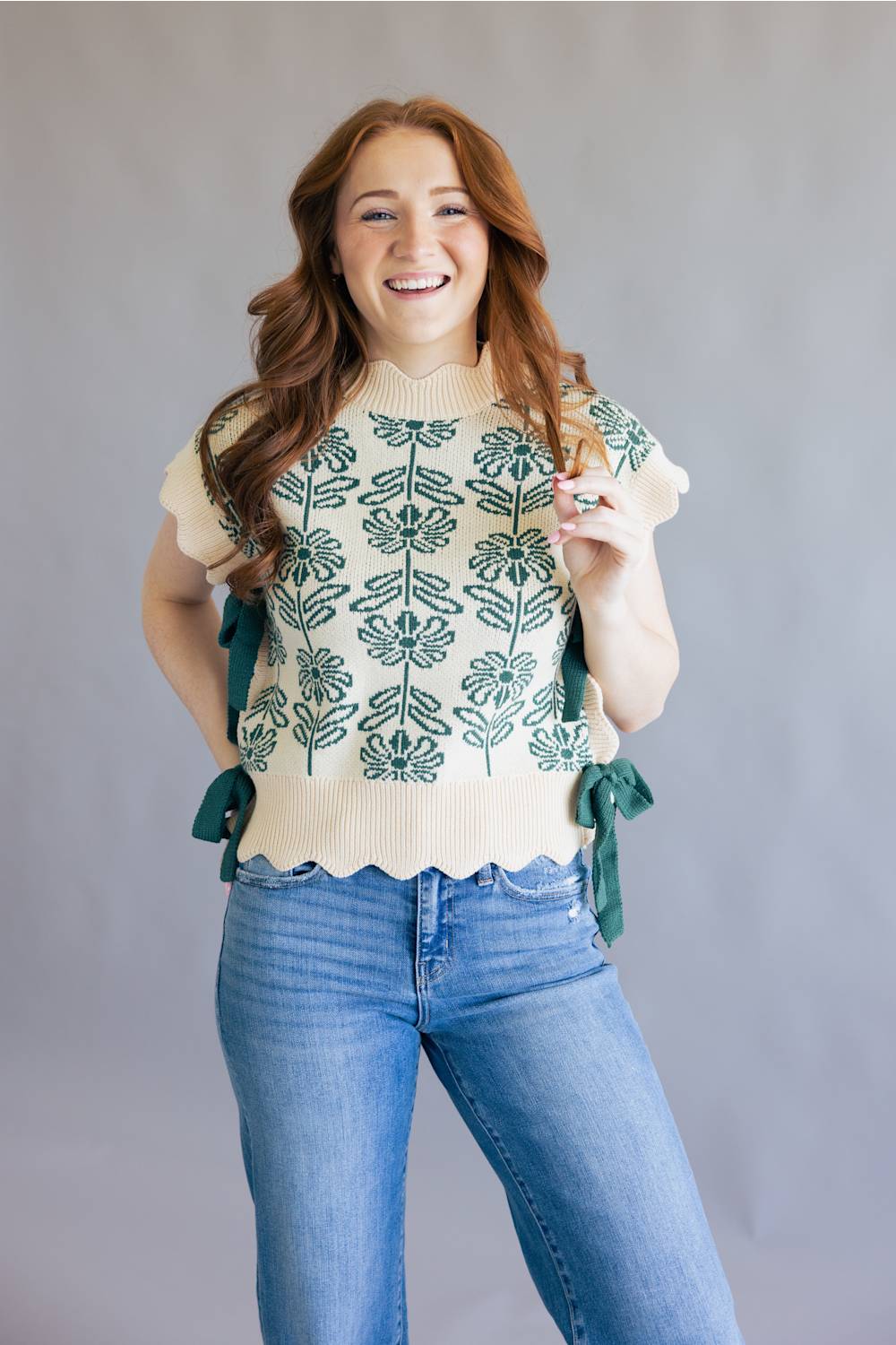 A smiling woman with long wavy red hair models a cream knit top with green floral embroidery and side ties, paired with blue jeans, against a plain gray studio background.