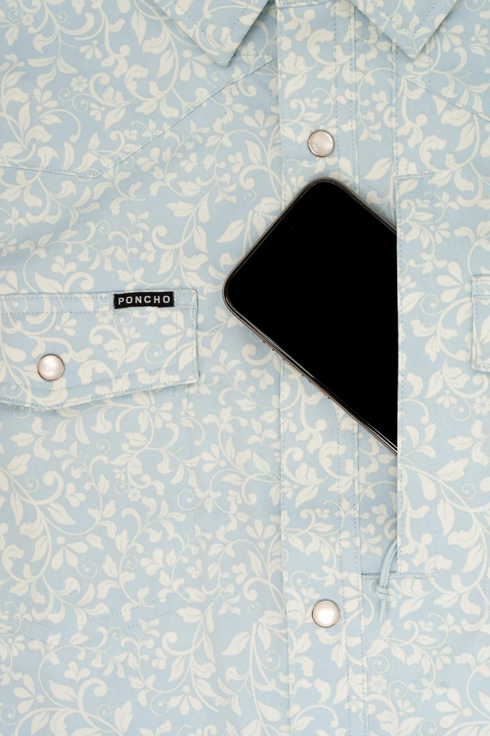 A close-up of a light blue and white damask-patterned button-down shirt by Poncho, featuring a chest pocket with silver snap buttons and a black smartphone tucked into the right pocket.