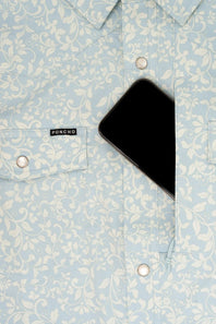 A close-up of a light blue and white damask-patterned button-down shirt by Poncho, featuring a chest pocket with silver snap buttons and a black smartphone tucked into the right pocket.