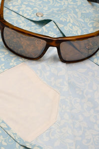 Brown tortoiseshell sunglasses with dark tinted lenses resting on a light blue and white patterned shirt with a white pocket.