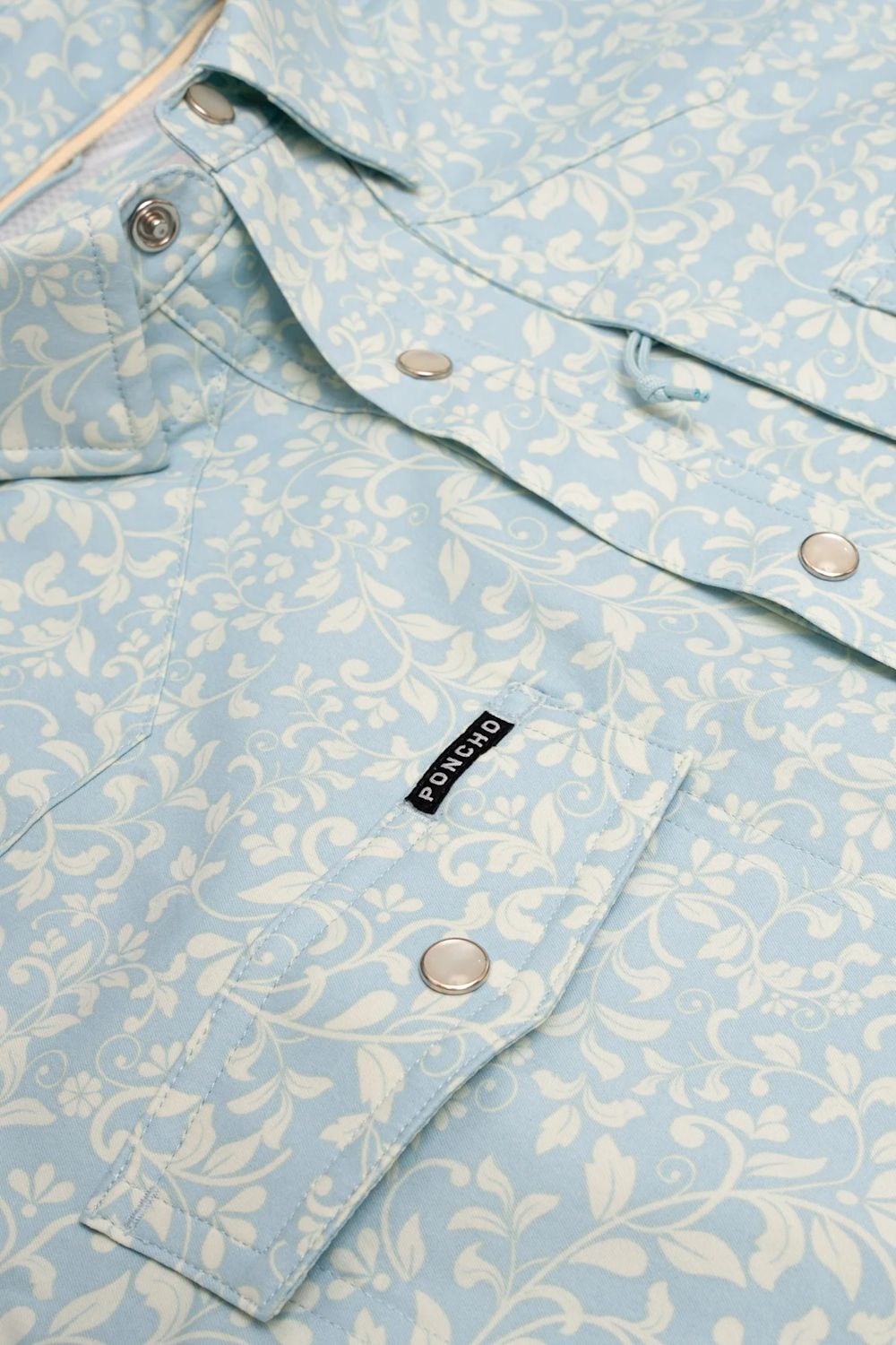 A close-up of a light blue and white floral patterned button-up shirt by Port & Company, featuring a chest pocket with a black brand label.