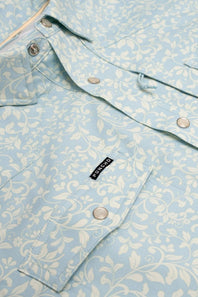 A close-up of a light blue and white floral patterned button-up shirt by Port & Company, featuring a chest pocket with a black brand label.