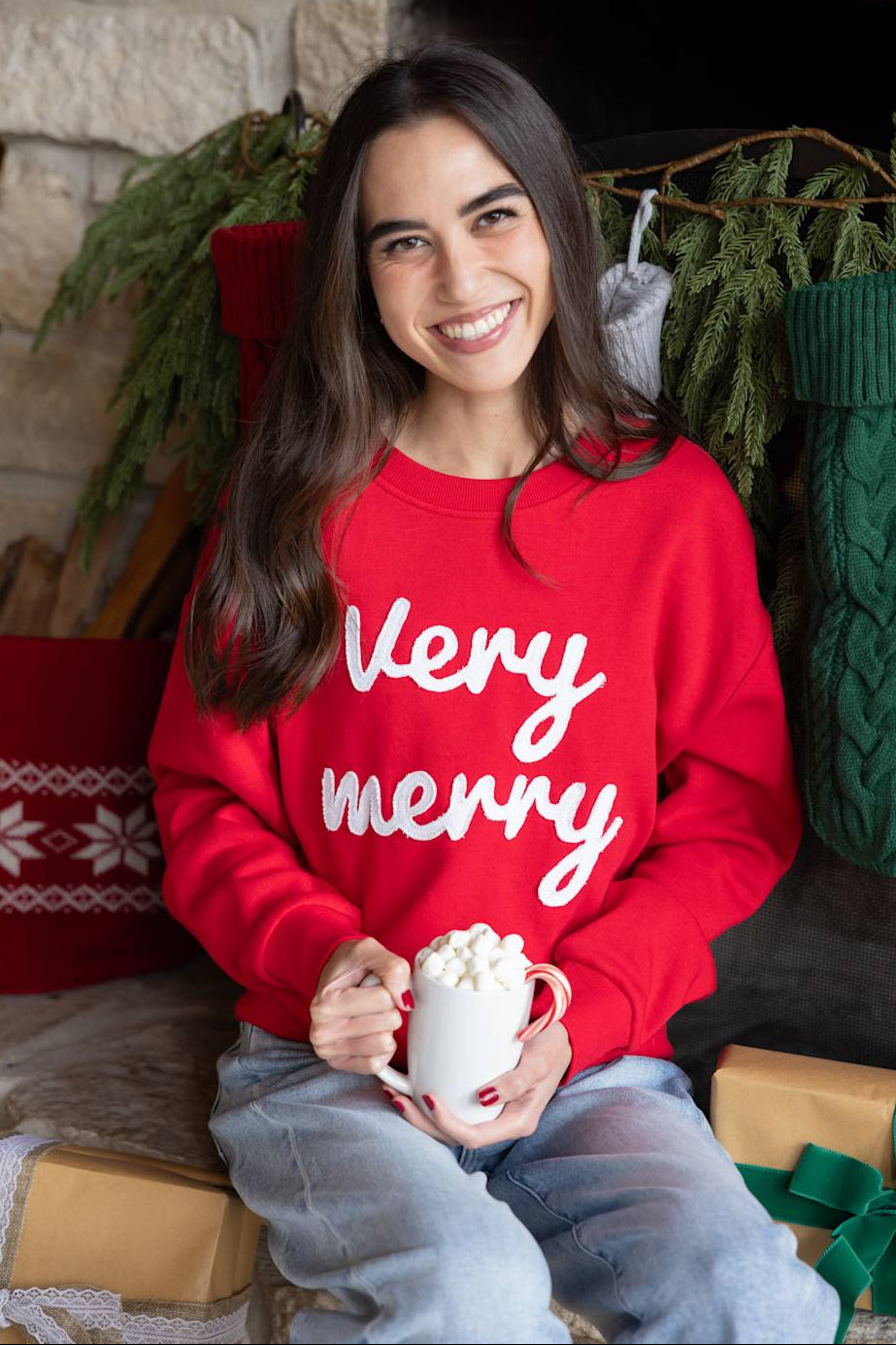 Holiday lifestyle photo of a dark-haired woman in a bright red "Very Merry" embroidered sweatshirt sitting near a stone fireplace with Christmas decorations.