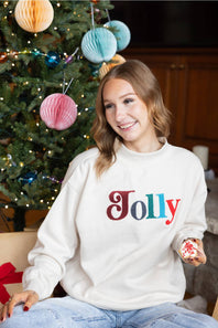 A smiling blond woman sitting next to a Christmas tree while holding a decorated cookie, wearing a beige mock-neck fleece pullover with multicolored chenille patch lettering that reads "Jolly" on the front.