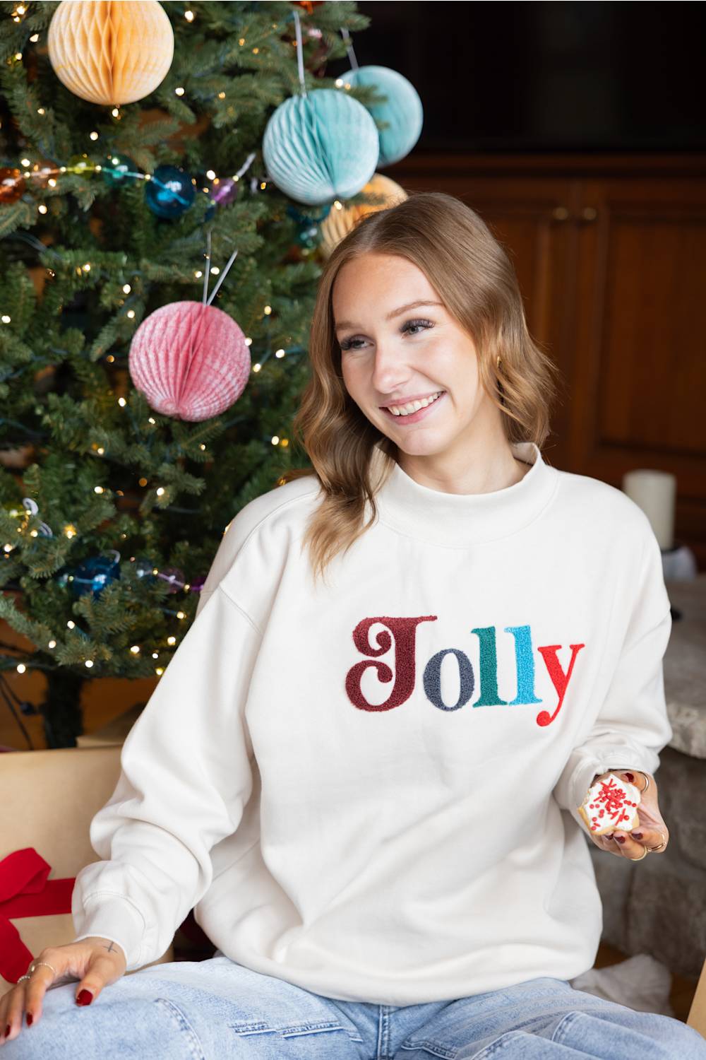 A smiling blond woman sitting next to a Christmas tree while holding a decorated cookie, wearing a beige mock-neck fleece pullover with multicolored chenille patch lettering that reads "Jolly" on the front.