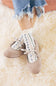 Top-down lifestyle view of a model wearing taupe suede Mary Jane ballet flats with ribbon tie lace-up straps, paired with floral patterned ruffled socks and cuffed frayed denim.