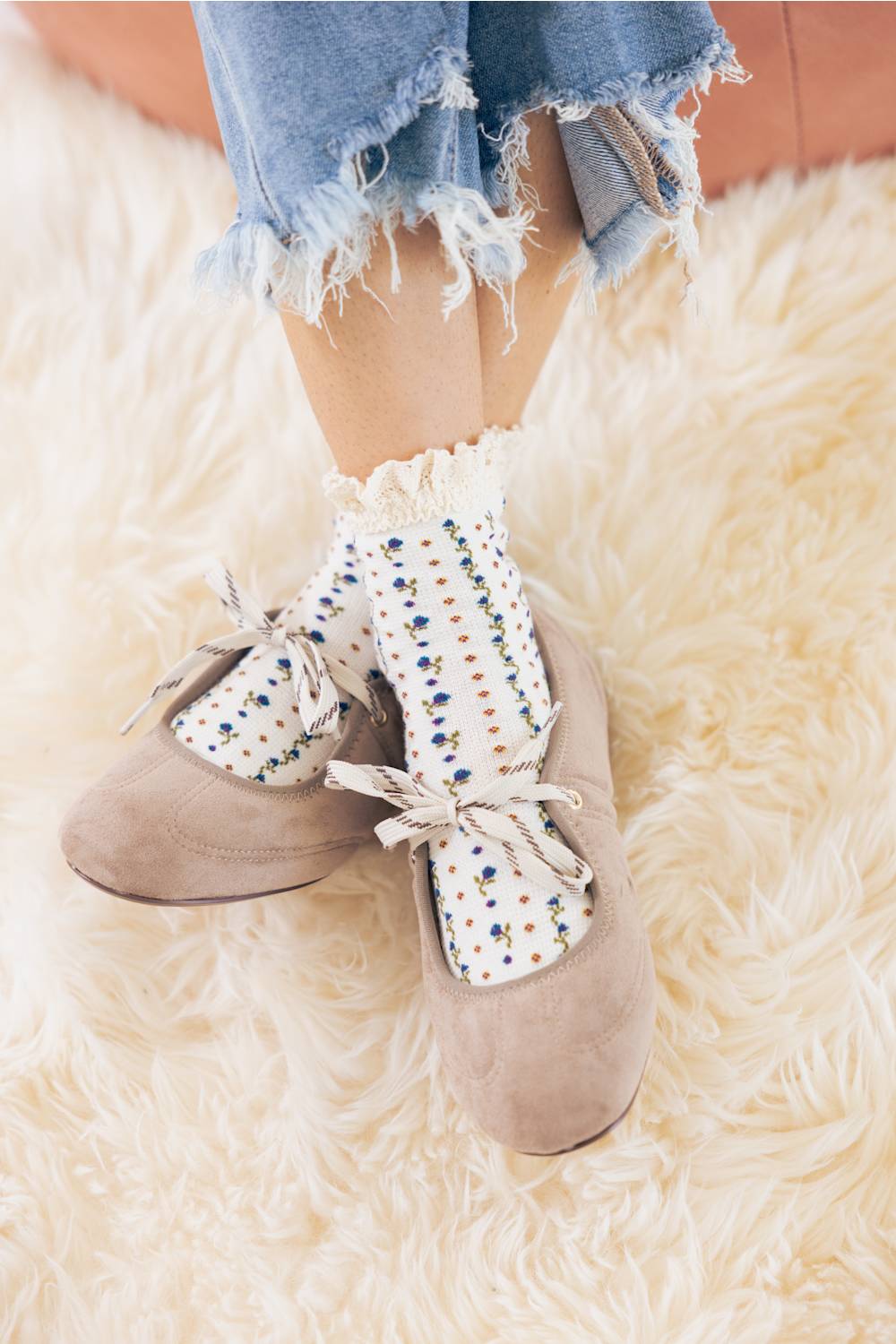 Top-down lifestyle view of a model wearing taupe suede Mary Jane ballet flats with ribbon tie lace-up straps, paired with floral patterned ruffled socks and cuffed frayed denim.