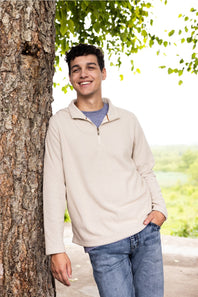 A young man smiling and leaning against a large tree trunk outdoors. He is wearing a light beige quarter-zip pullover sweater over a blue-gray t-shirt and blue jeans.