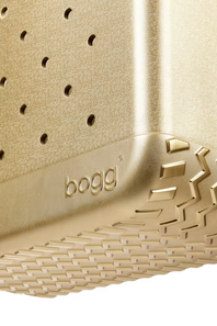 Close-up of a gold-colored Bogg brand product, showing a textured surface with a perforated top section, a treaded bottom edge, and the embossed 'bogg' logo.